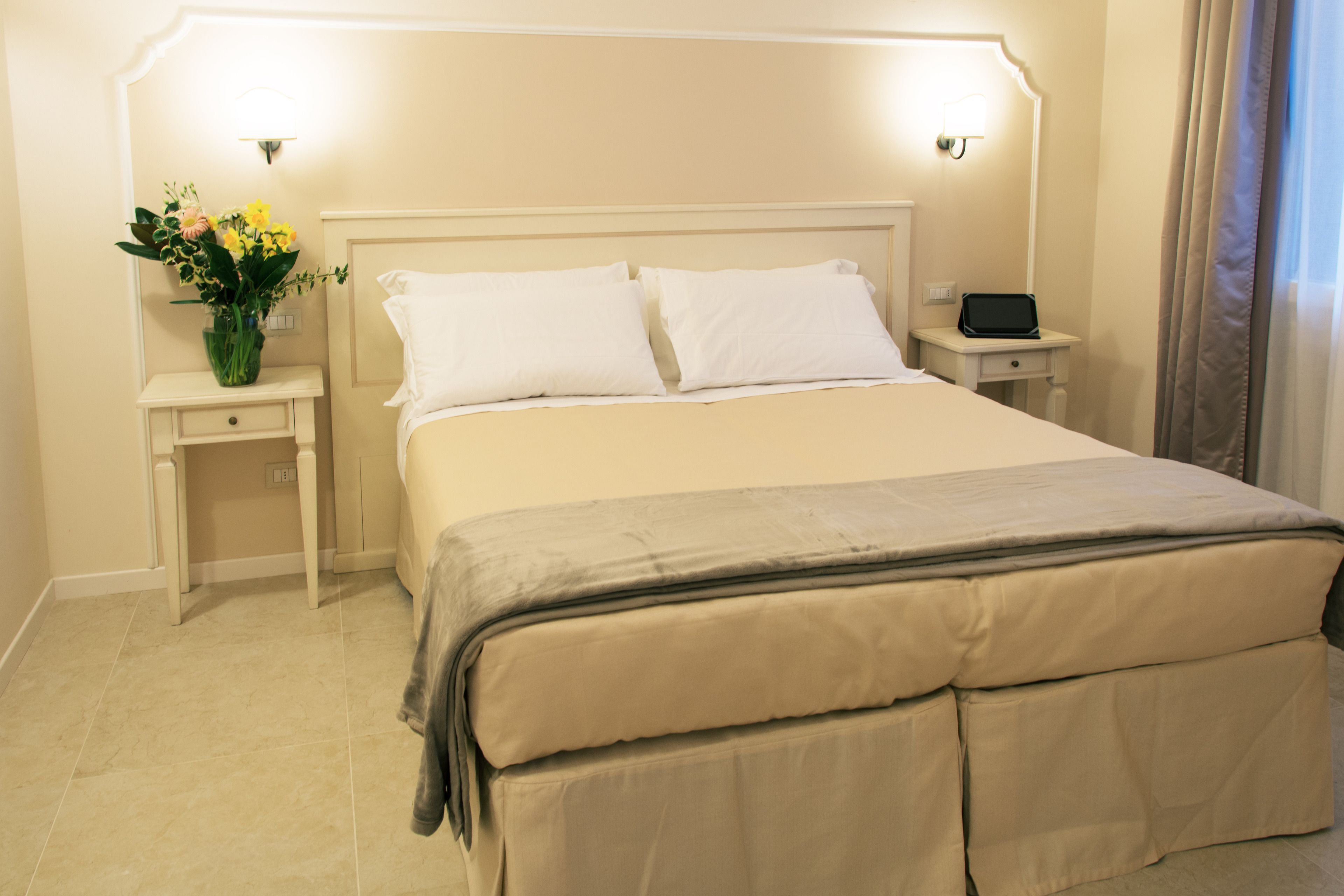 A neatly made bed with white linens, a bedside table with a lamp, and a vase of flowers.