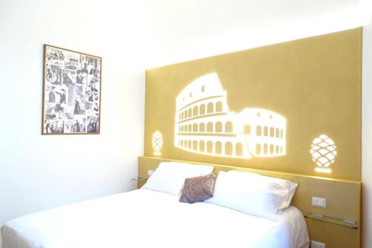 A hotel room with a bed, a framed photo collage on the wall, and a large illuminated image of the Colosseum.