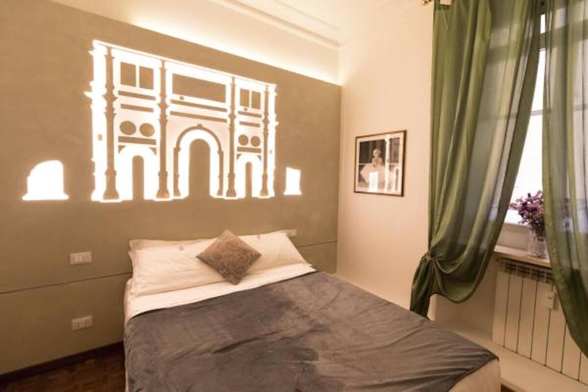 A bedroom with a bed, a wall-mounted projector displaying a building silhouette, a window with green curtains, and a framed picture on the wall.