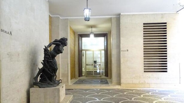 Un couloir orné d’une sculpture, doté d’un sol à motifs, d’une porte portant l’inscription « ESCALIER A » et d’une grille d’aération sur le mur.