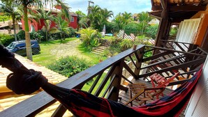 A hammock on a wooden deck with a view of a garden and a red building. - Pousada Vistacalma (Imbituba)