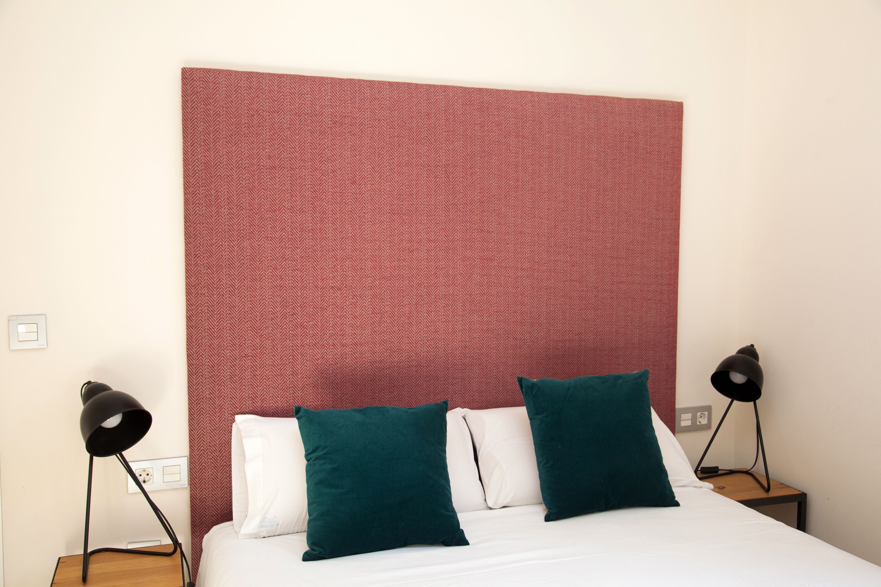 A hotel room with a bed, two green pillows, a white pillow, a red textured headboard, and two black floor lamps.