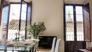 A dining area with a glass table, chairs, and a TV. There are windows with open shutters, and a view of a building with a tiled roof.