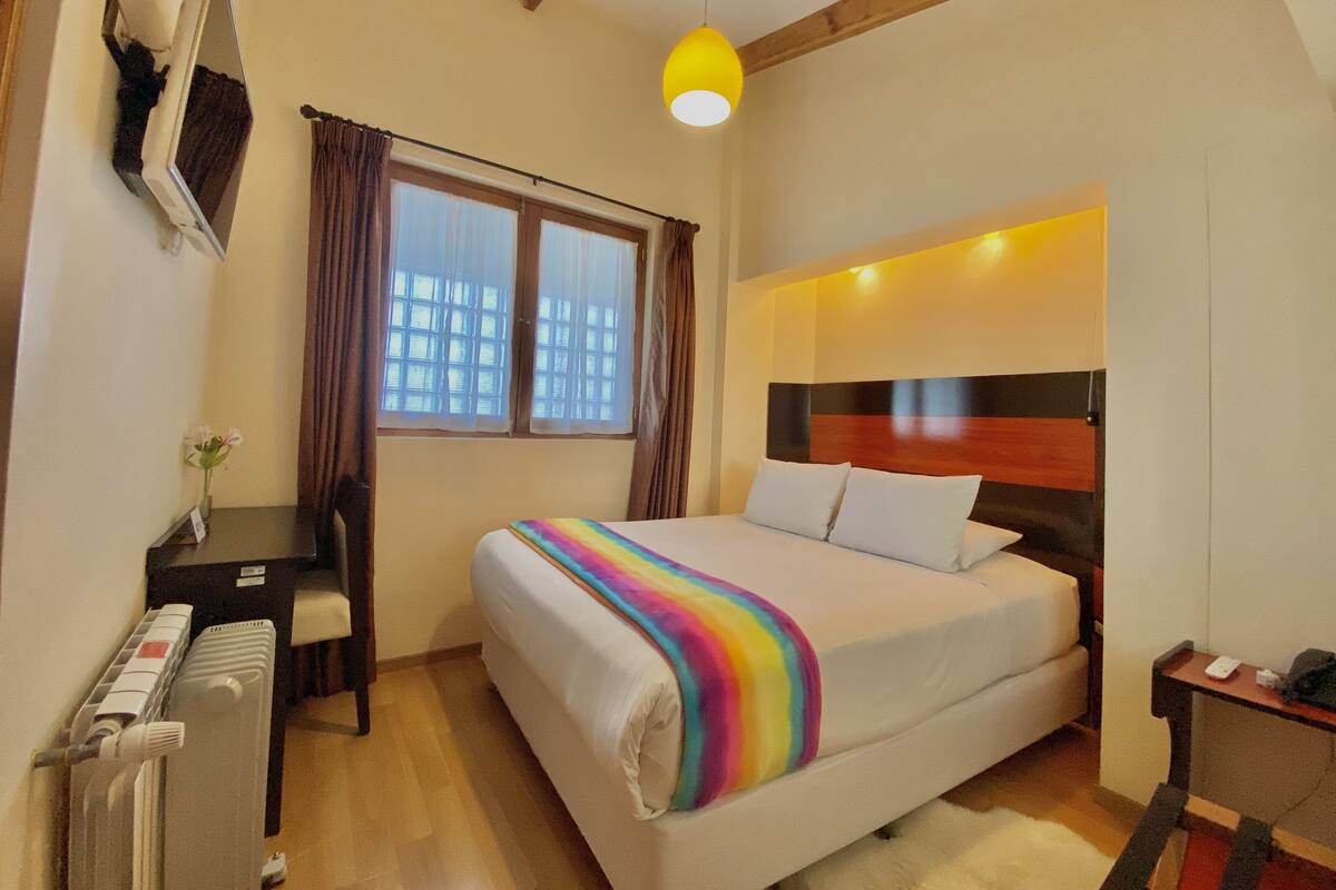 Un dormitorio con cama, escritorio, silla, radiador y ventana con cortinas.