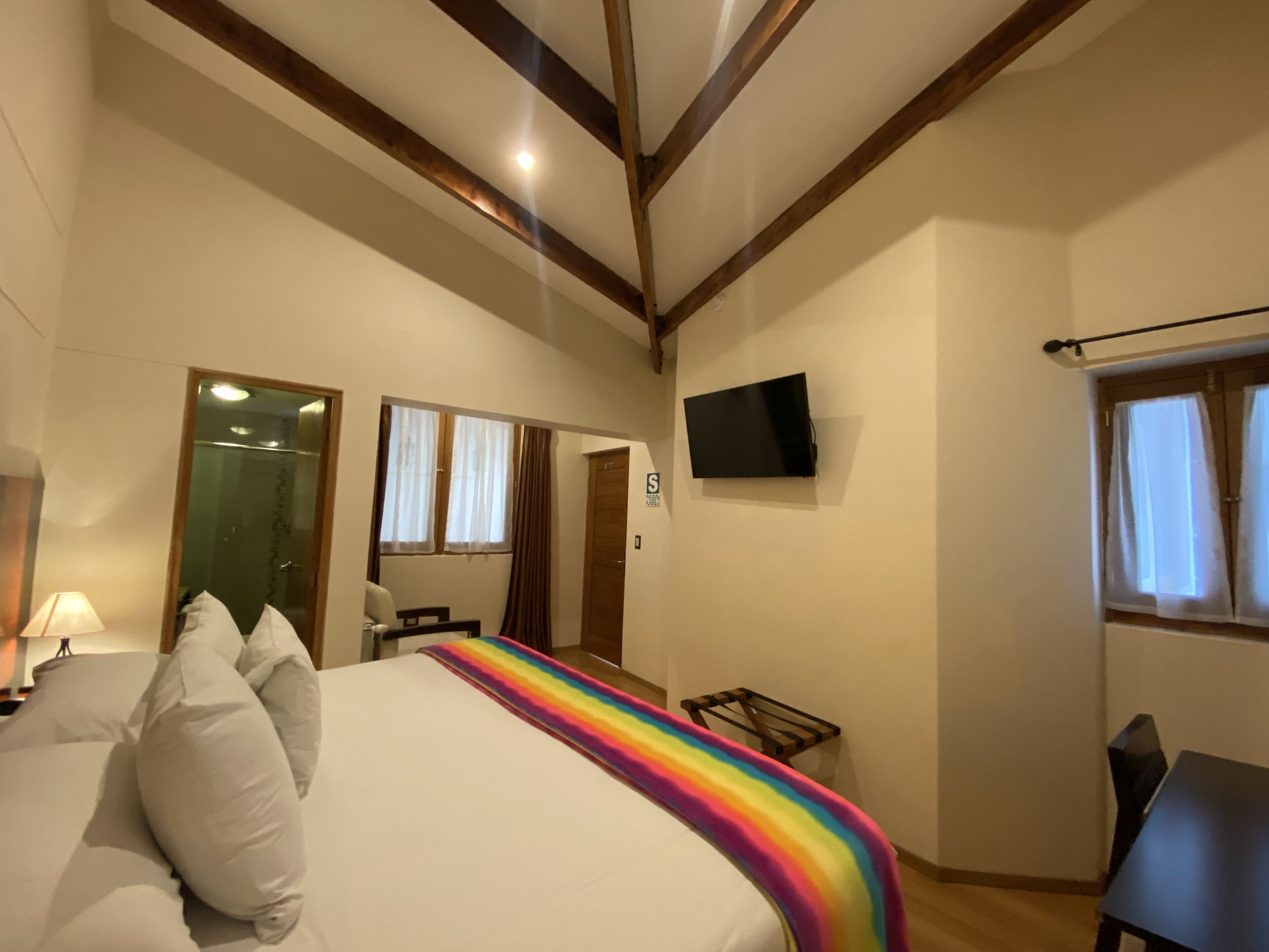 Un dormitorio con una cama, un televisor montado en la pared, una ventana con cortinas y una puerta.
