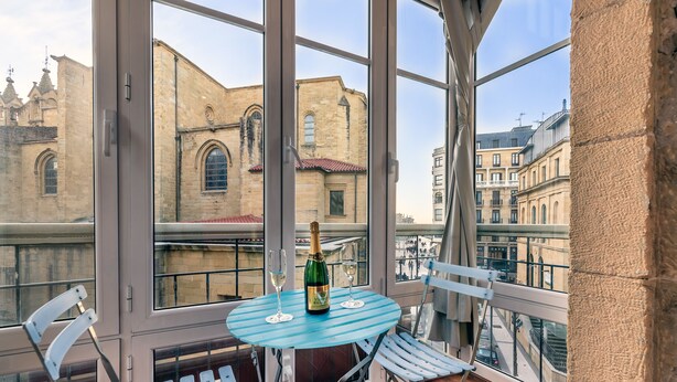 A balcony with a table and chairs, a bottle of champagne, and a view of historic buildings. - Pensión Ab Domini (San Sebastián)