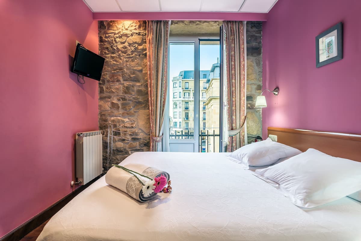 A hotel room with a bed, a TV mounted on the wall, a window with curtains, and a stone wall.
