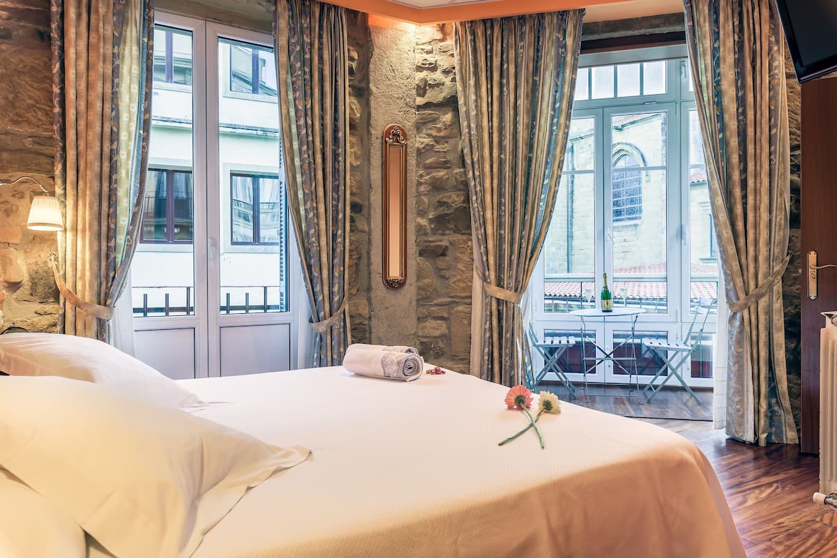 A hotel room with a stone wall, large windows with curtains, a bed with white linens, and a balcony with outdoor furniture.