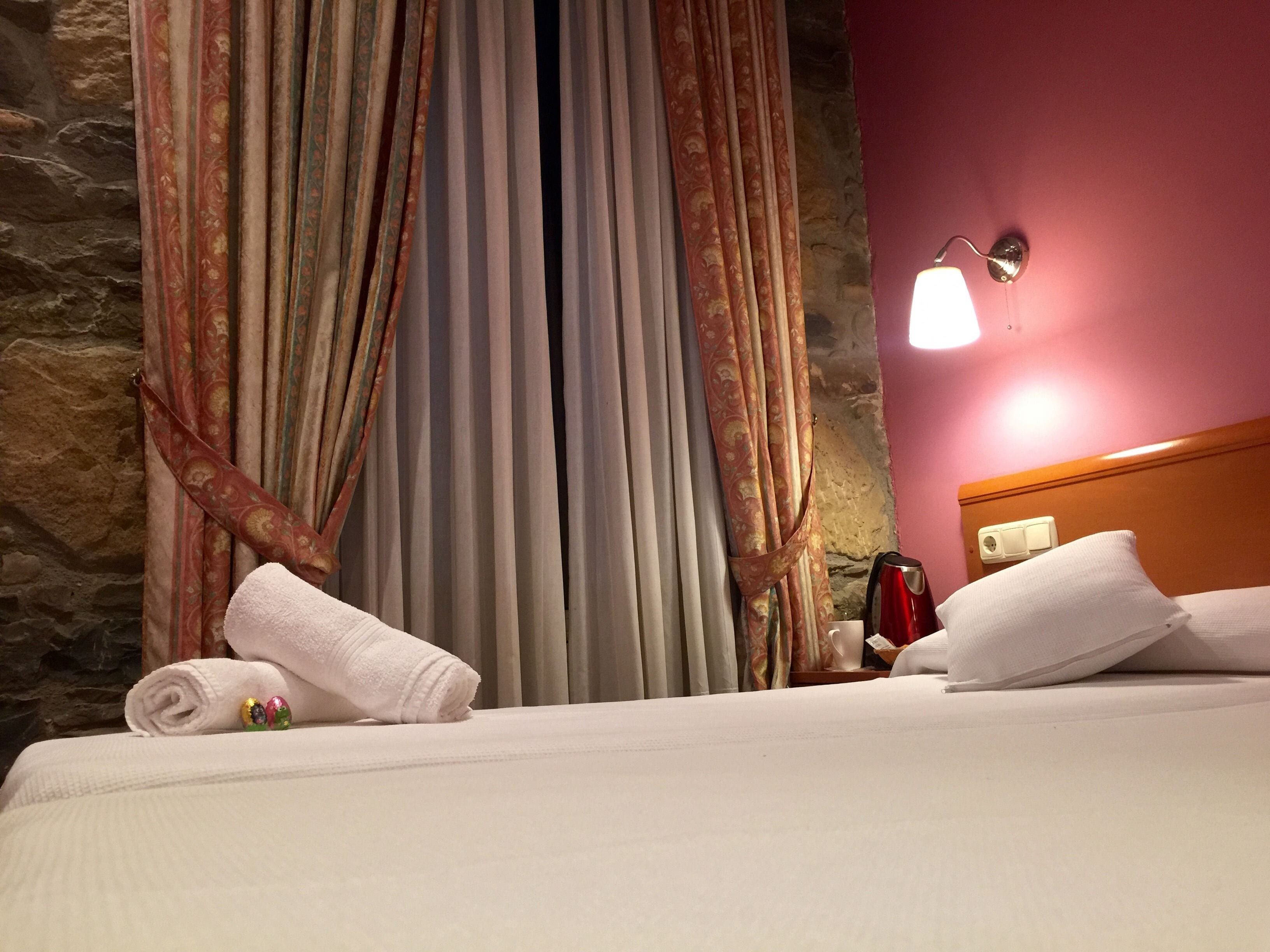 Habitación de hotel con cama, una toalla enrollada, una tetera roja y una mesita de noche.