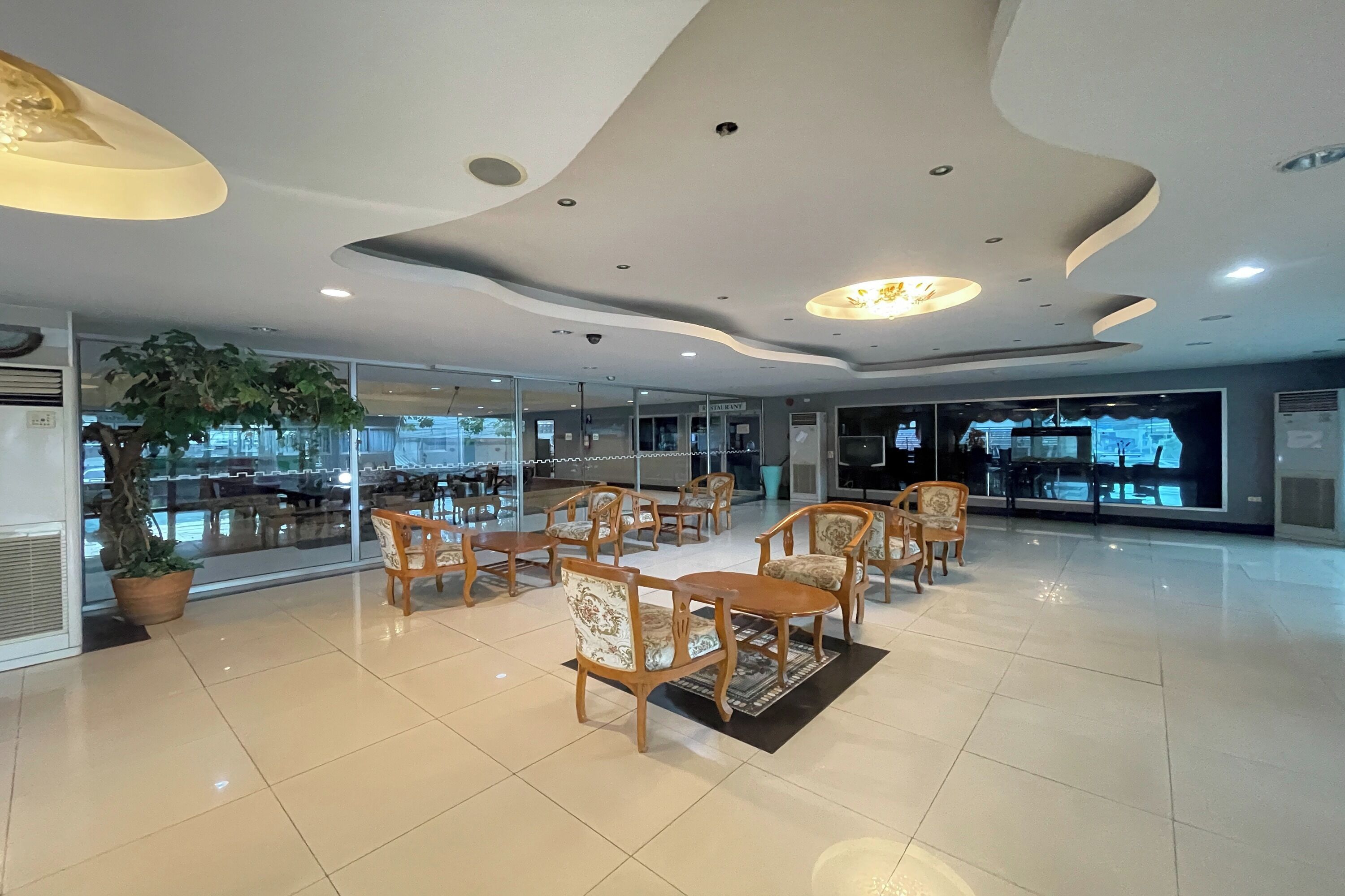 A spacious lobby with a modern ceiling design, a large potted plant, and a seating area with wooden chairs and tables.