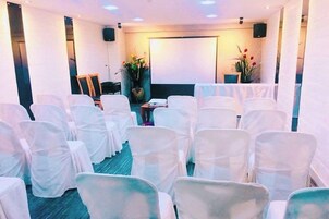 A conference room with rows of white chairs, a projector screen, and a speaker on the wall.