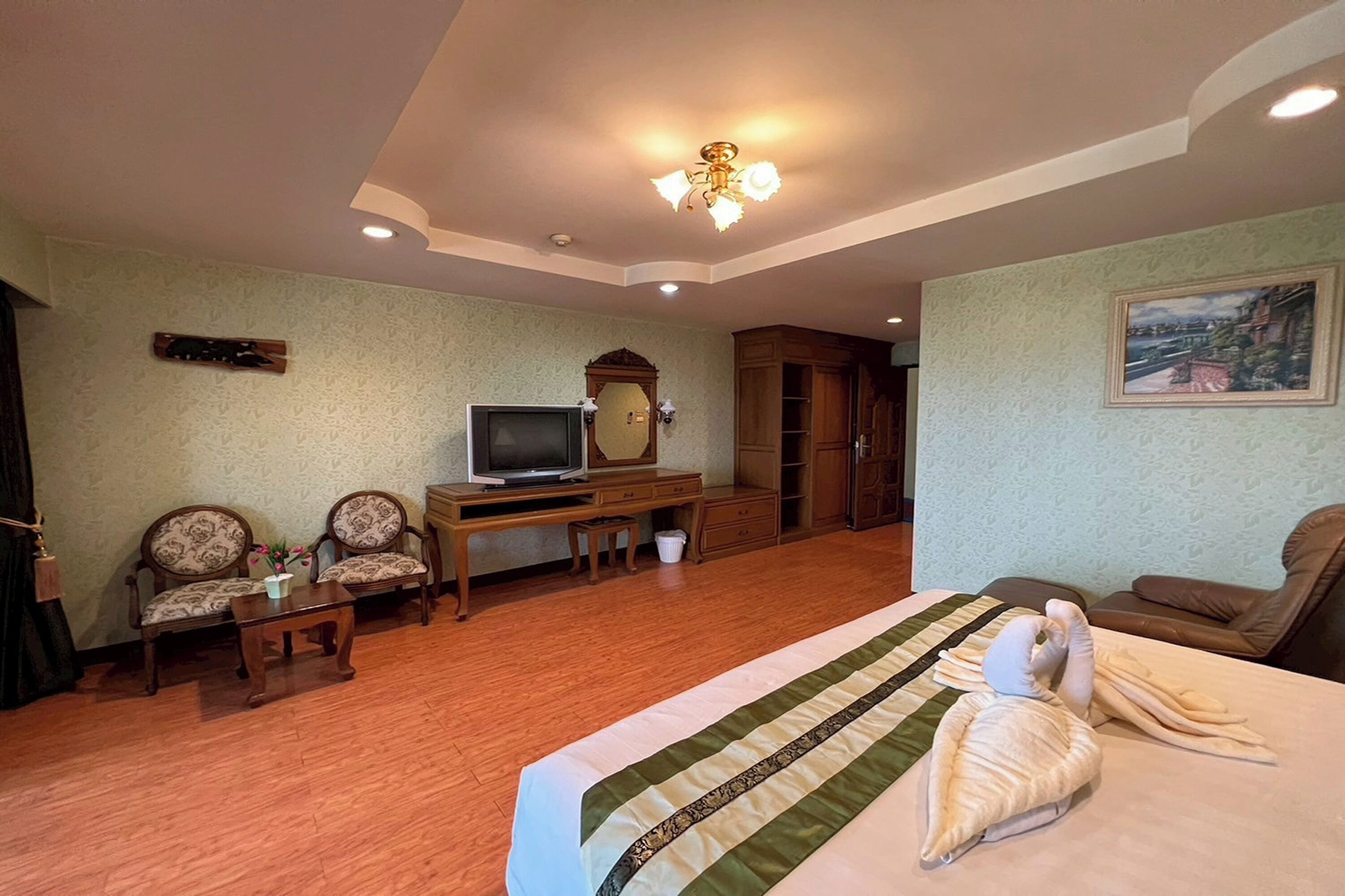 Habitación de hotel con una cama, dos sillones, un escritorio con televisor, un armario de madera y un cuadro en la pared.