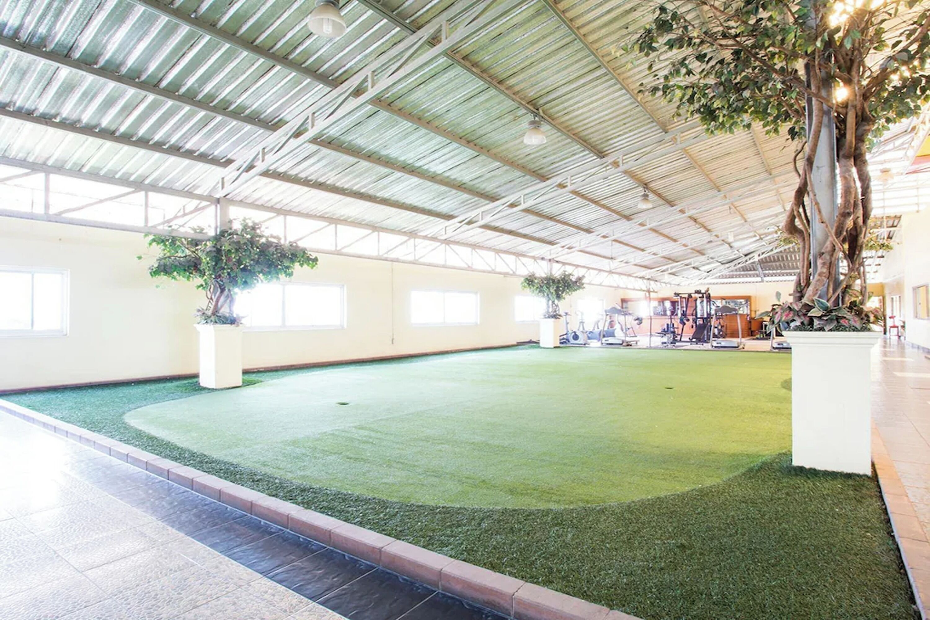 Amplo espaço interno com grama sintética verde, plantas grandes em vasos e teto alto com vigas de metal.