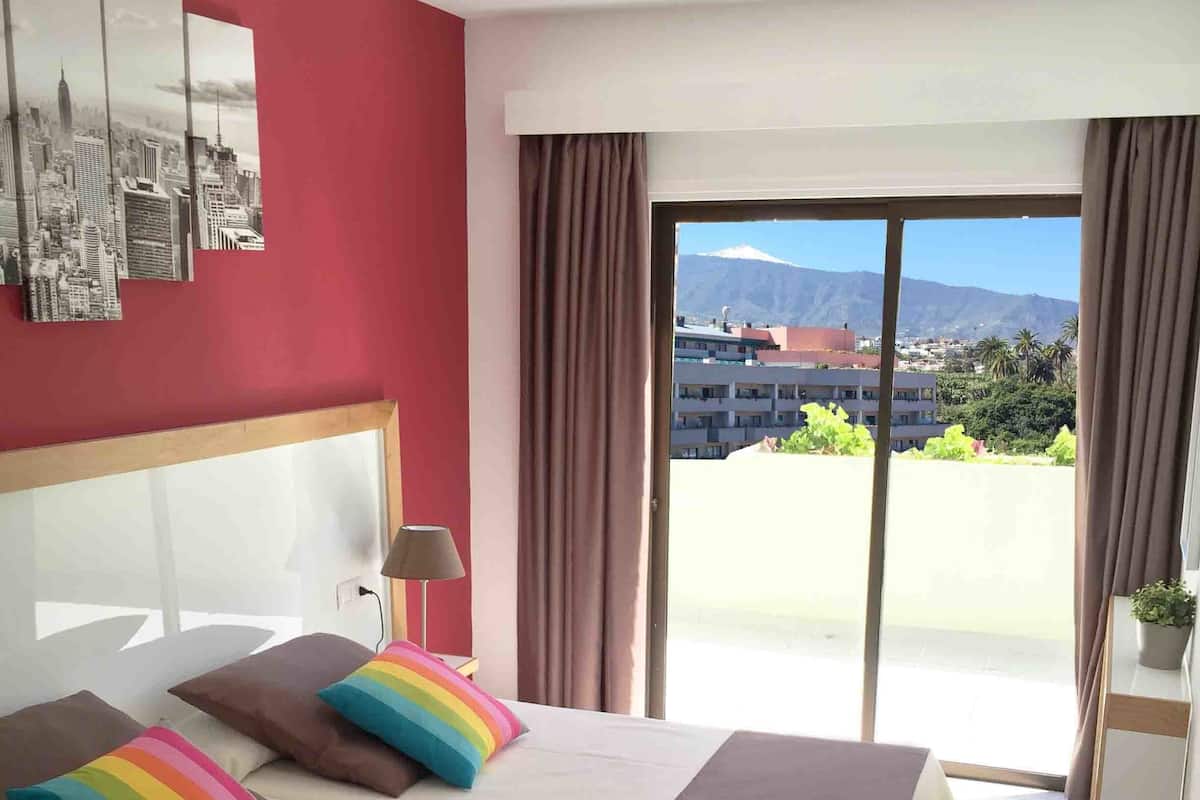 Une chambre d’hôtel avec un lit, une œuvre d’art représentant un paysage urbain accrochée au mur, une vue sur les bâtiments et les montagnes par la fenêtre, et un balcon avec des rideaux.