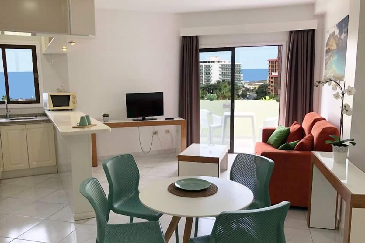 Un appartement moderne comprenant une cuisine, un coin repas et un salon. Il dispose d’une grande fenêtre offrant une vue sur la mer et les bâtiments environnants.