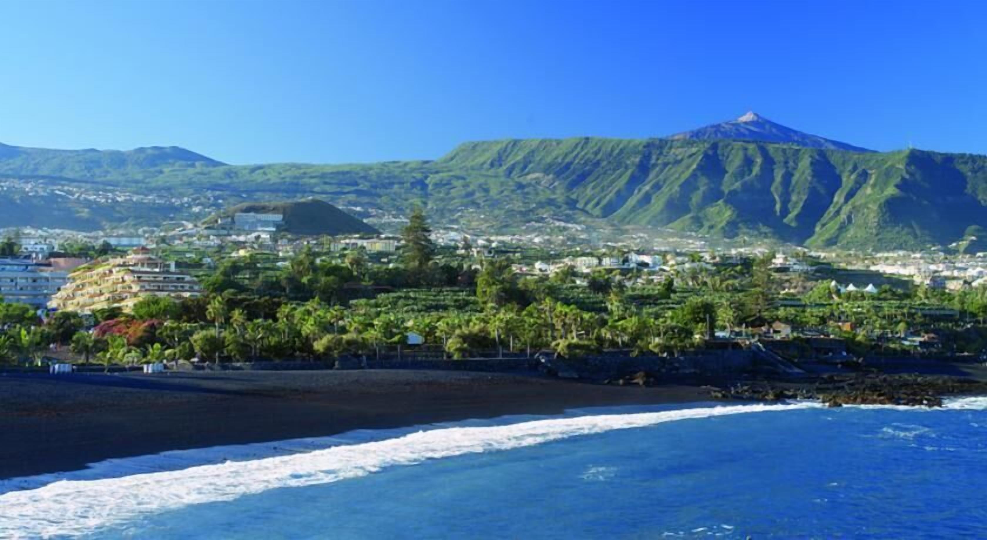 Un pueblo costero con una montaña destacada al fondo, una playa y una combinación de edificios residenciales y comerciales.