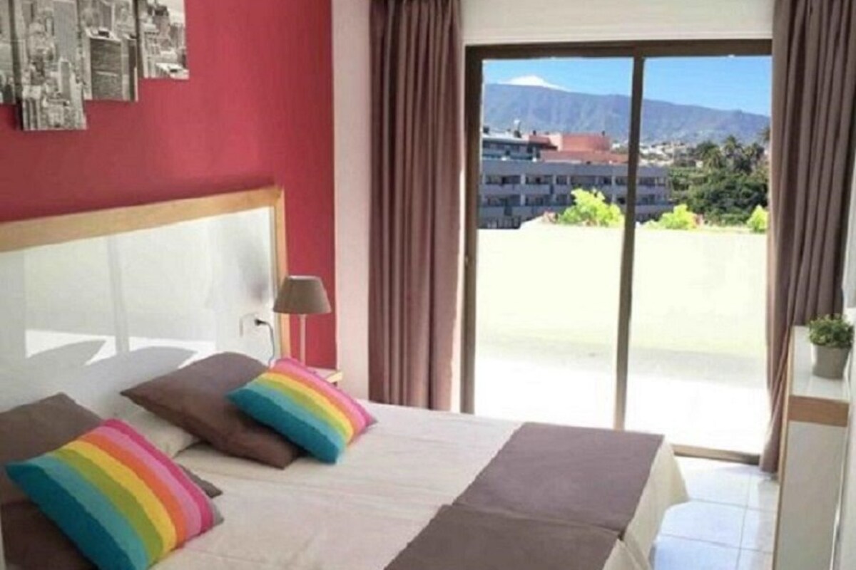 Hotelkamer met een bed, een hoofdeinde, een raam met gordijnen en uitzicht op een stadsbeeld en bergen.