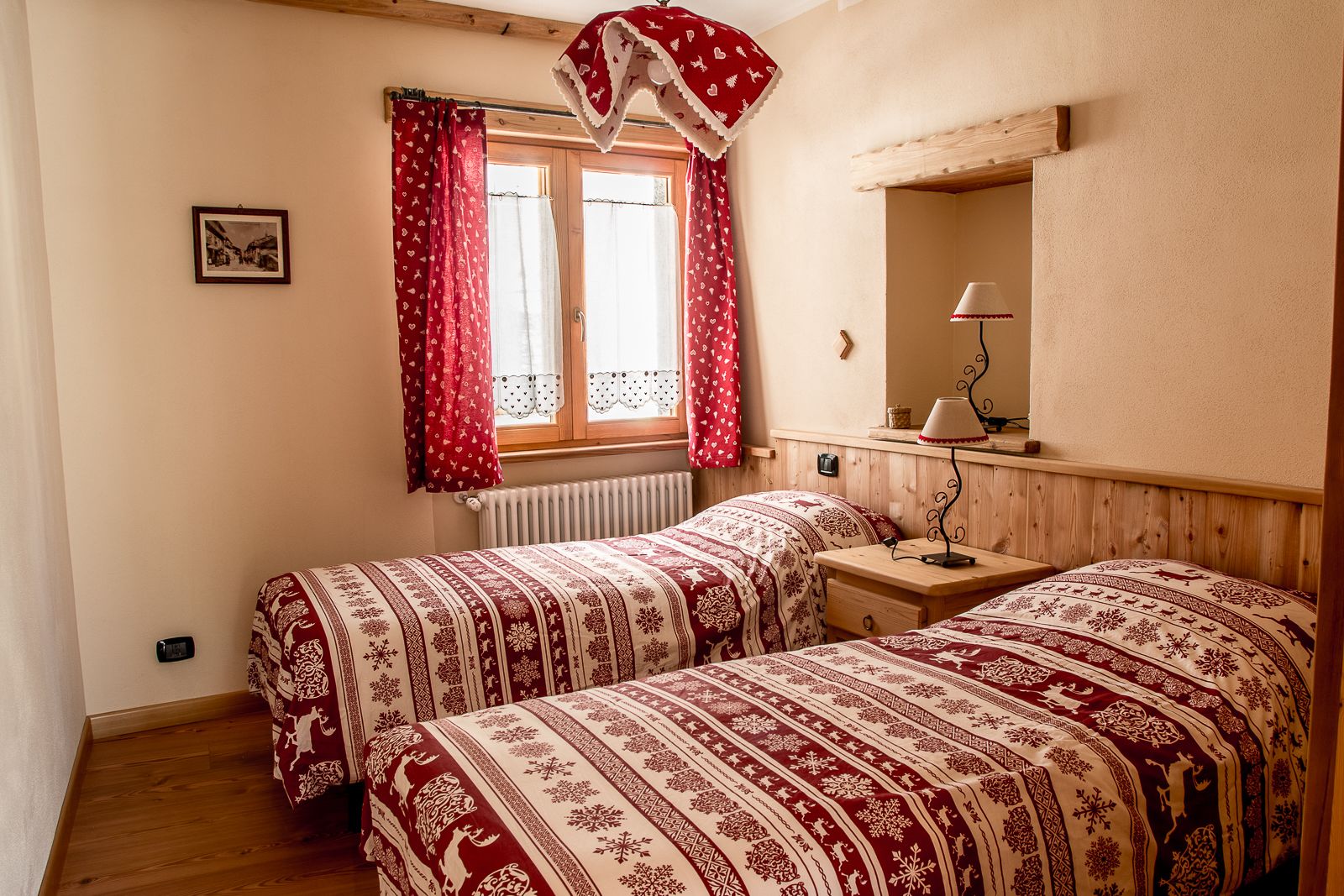 A room with two beds, a window with red curtains, a bedside table with a lamp, and a framed picture on the wall.