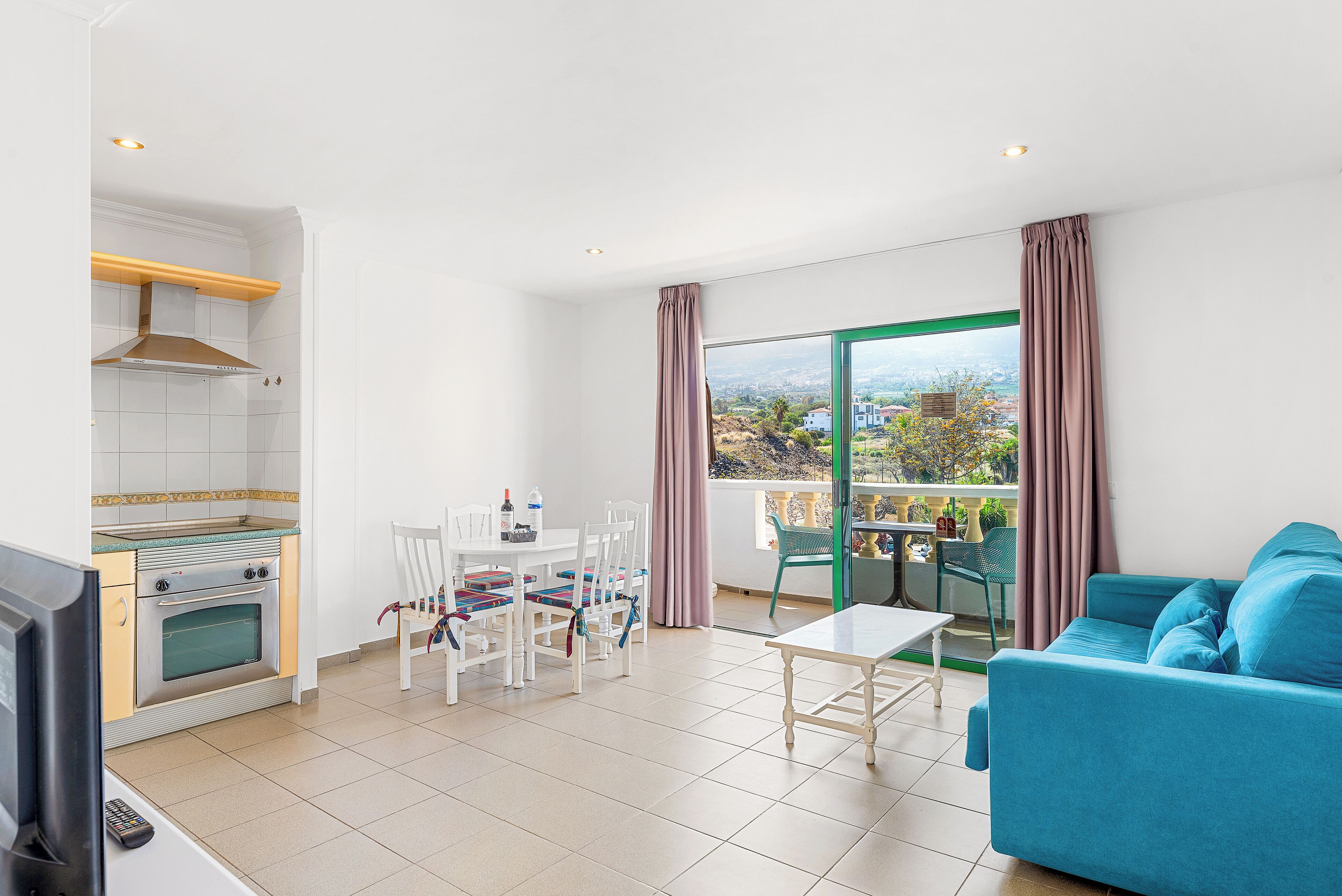 A modern kitchen with a built-in oven, a dining table with chairs, a blue sofa, and a balcony with a view.