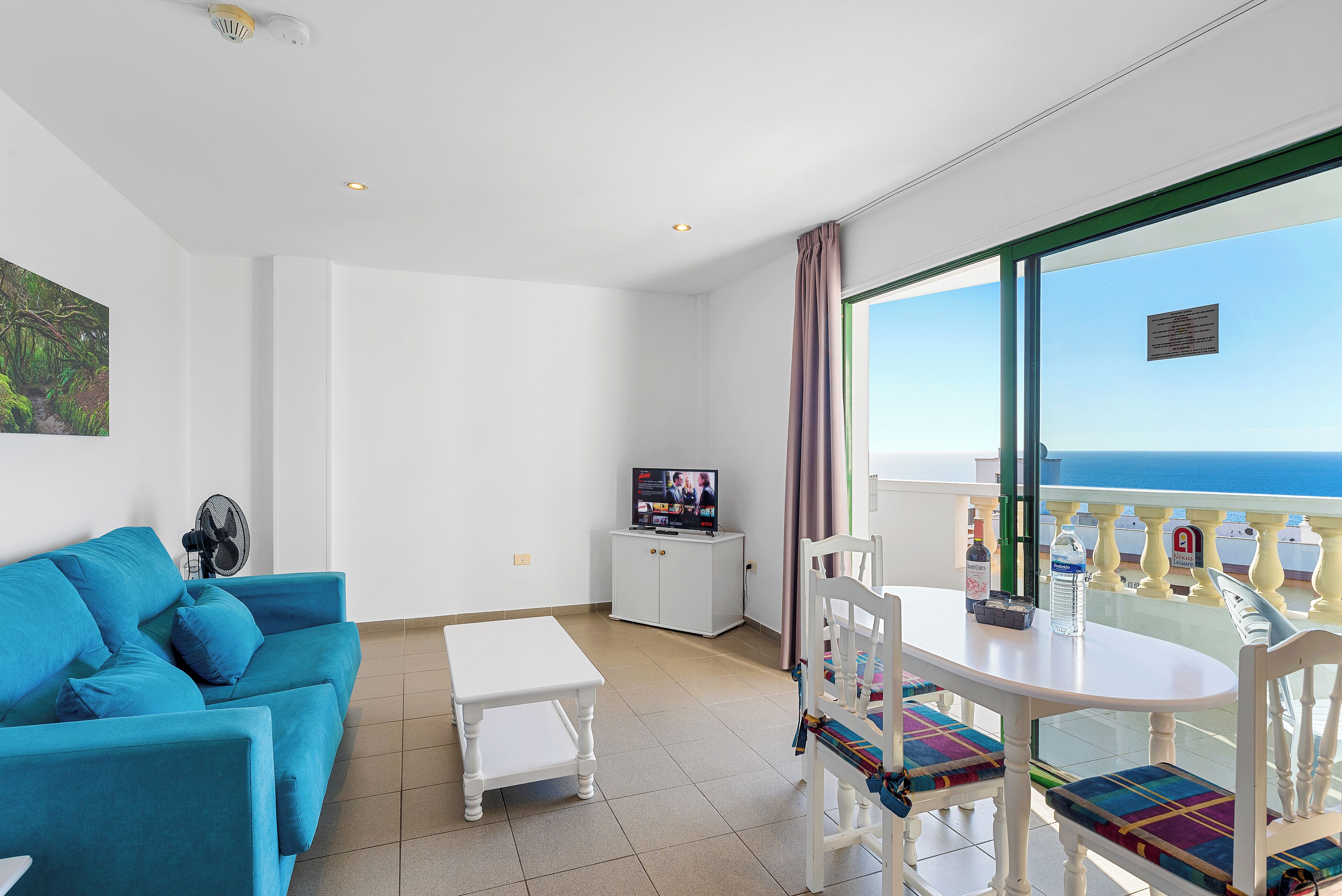 A room with a blue sofa, a small table, a TV on a cabinet, and a balcony with a view of the sea.
