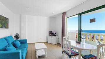 A room with a blue sofa, a small table, a TV on a cabinet, and a balcony with a view of the sea.