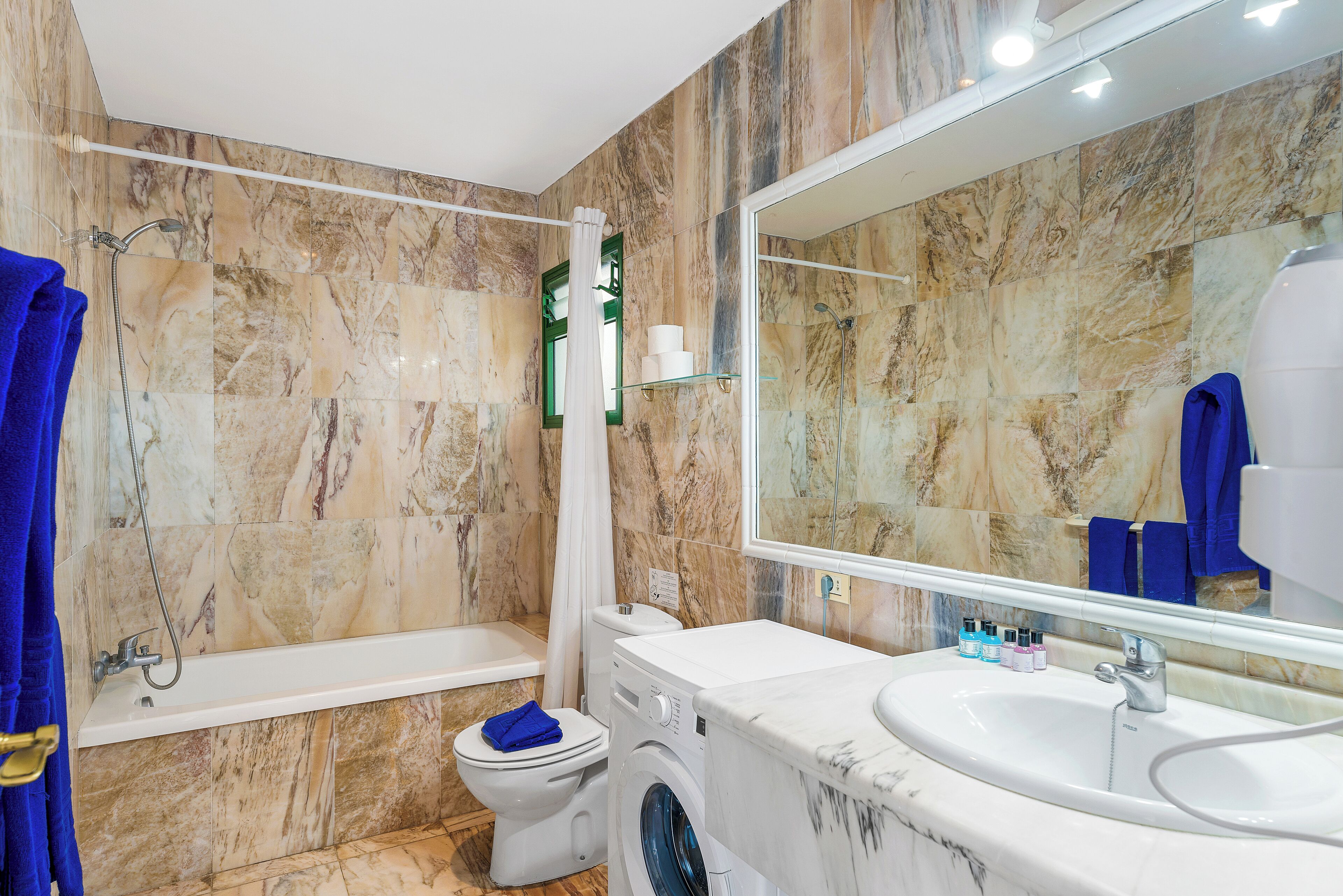 A bathroom with a marble wall, a bathtub, a toilet, a washing machine, and a sink.