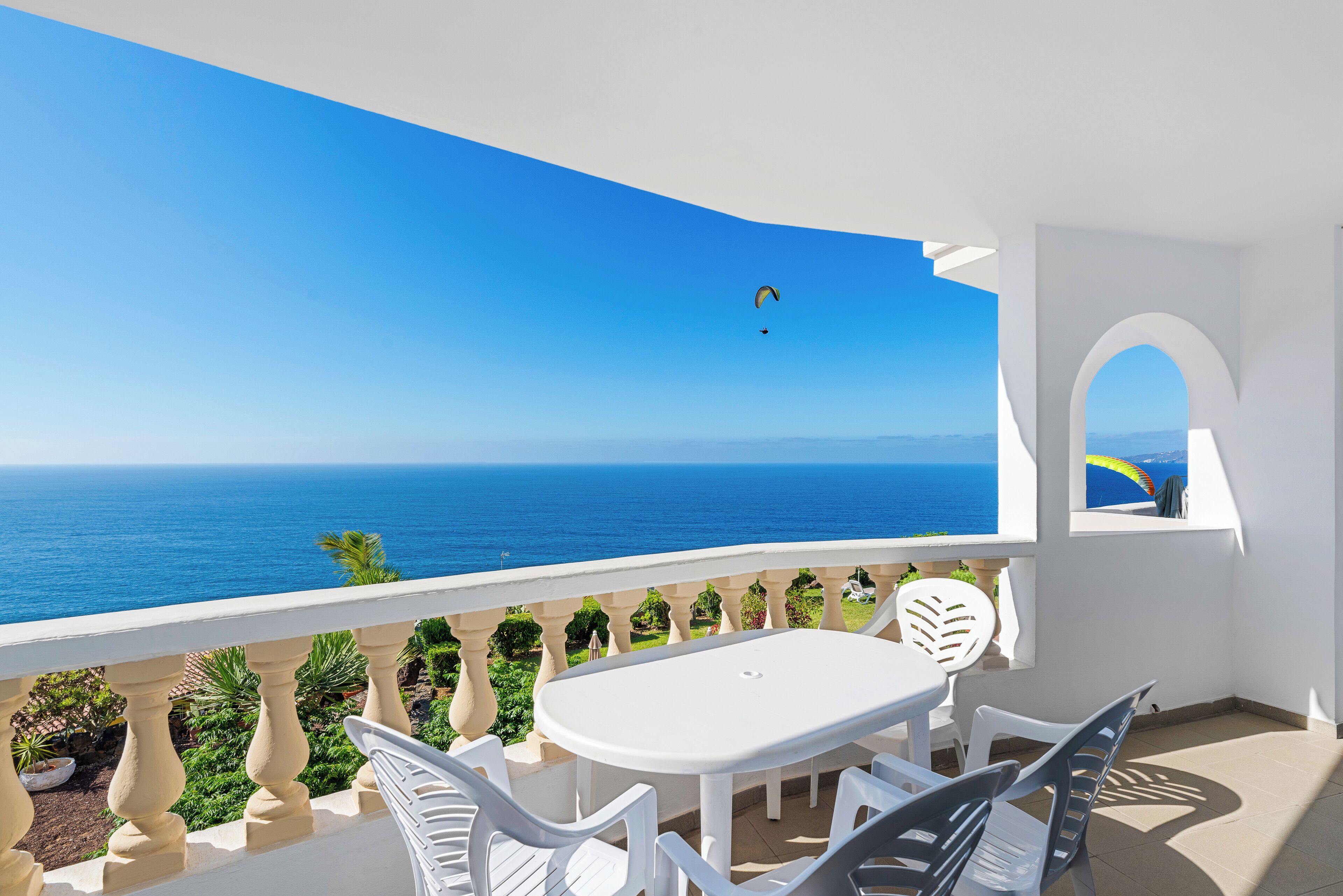 Varanda com mesa e cadeiras brancas, com vista para um mar azul e uma ilha ao longe.