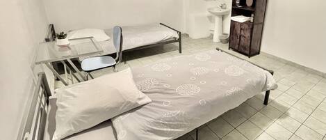Una stanza piccola e pulita con due letti singoli, un comodino, uno specchio, una finestra, un lavandino e un dipinto raffigurante Buddha.