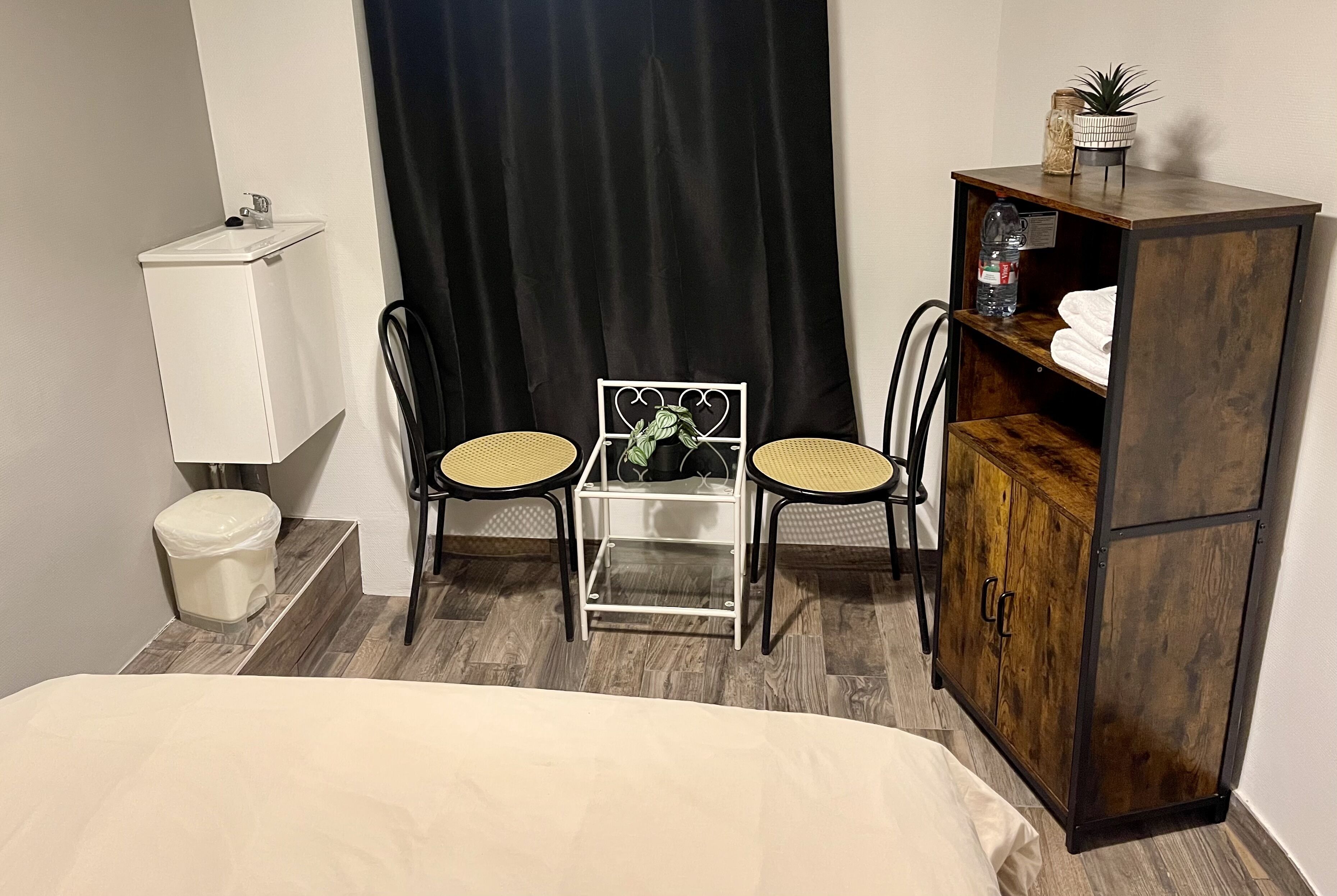 A small room with a bed, a white sink, two wicker chairs, a small table with a plant, and a wooden cabinet.