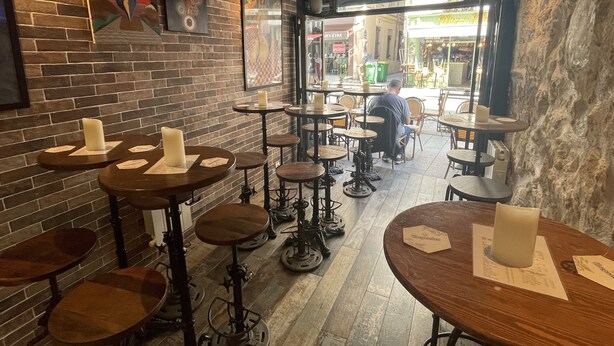 A rustic interior with brick walls, wooden tables, and stools. There are paintings on the walls and a man sitting at a table. - Le Globe (Paris)
