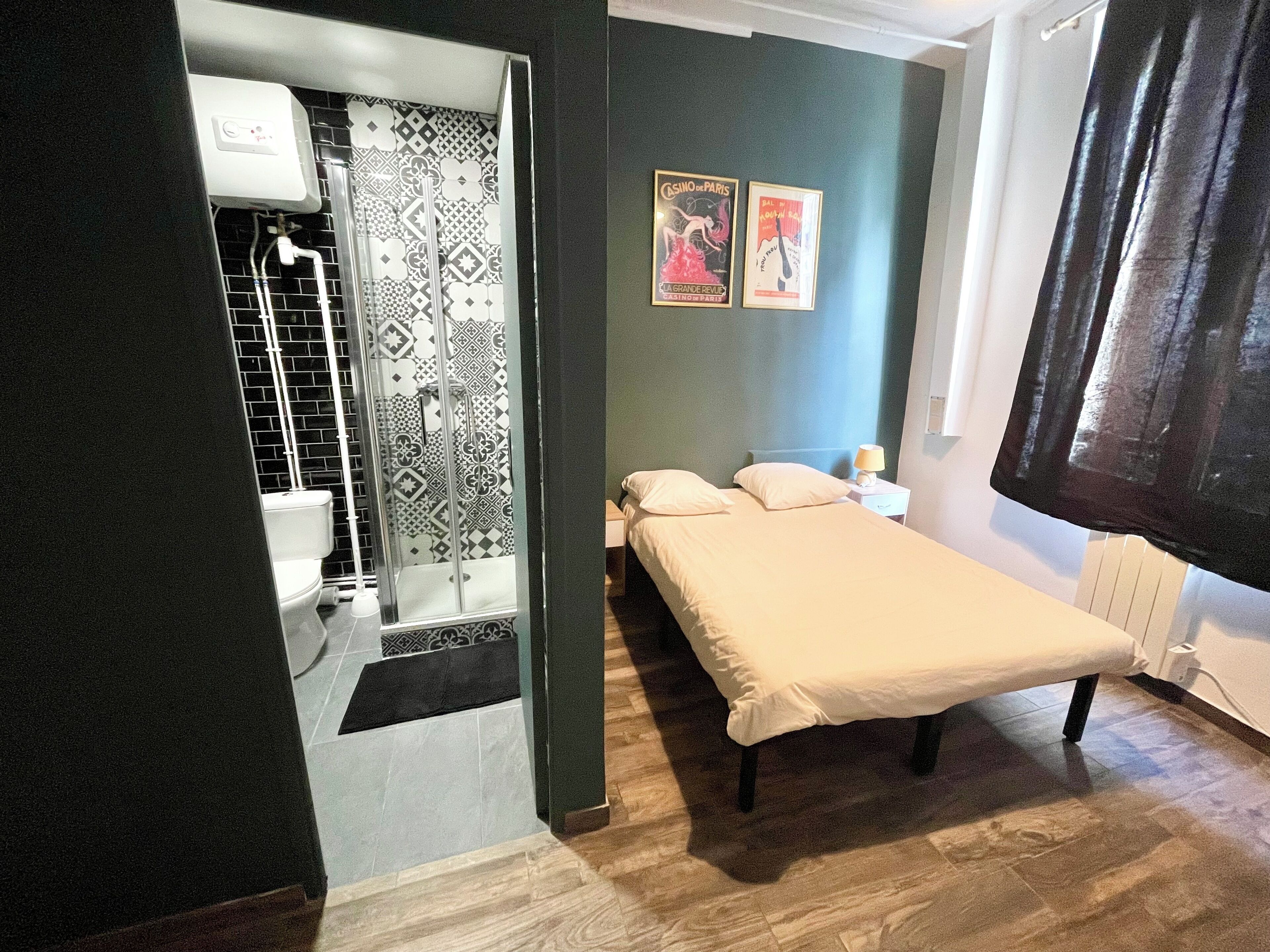 A bedroom with a bed, a nightstand, a lamp, a shower with a patterned wall, and two posters on the wall.