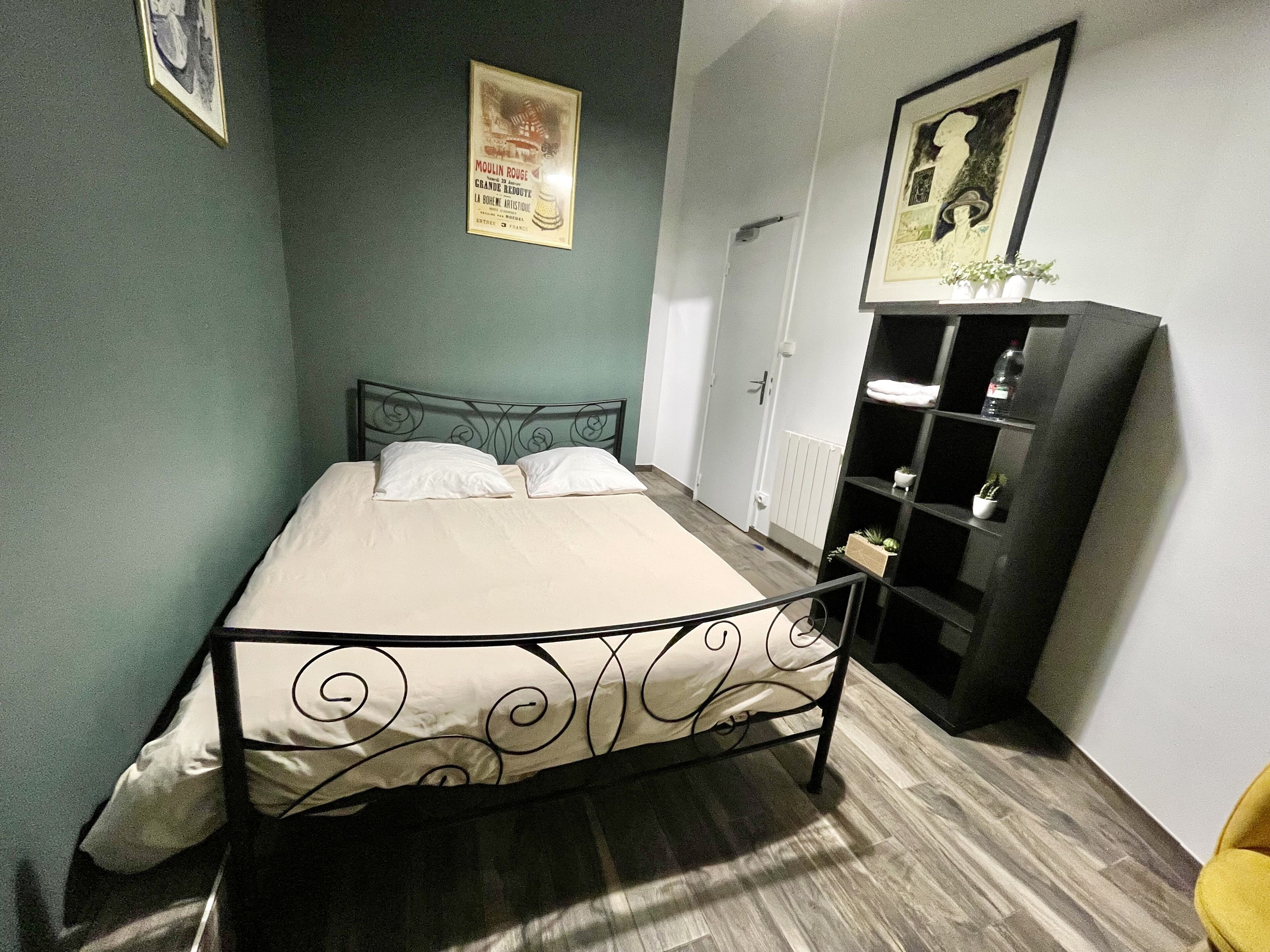 Ein ordentlich bezogenes Bett mit Metallgestell und dekorativem Kopfteil, ein kleiner Nachttisch mit Lampe sowie ein an der Wand befestigtes Regal mit einer Topfpflanze.