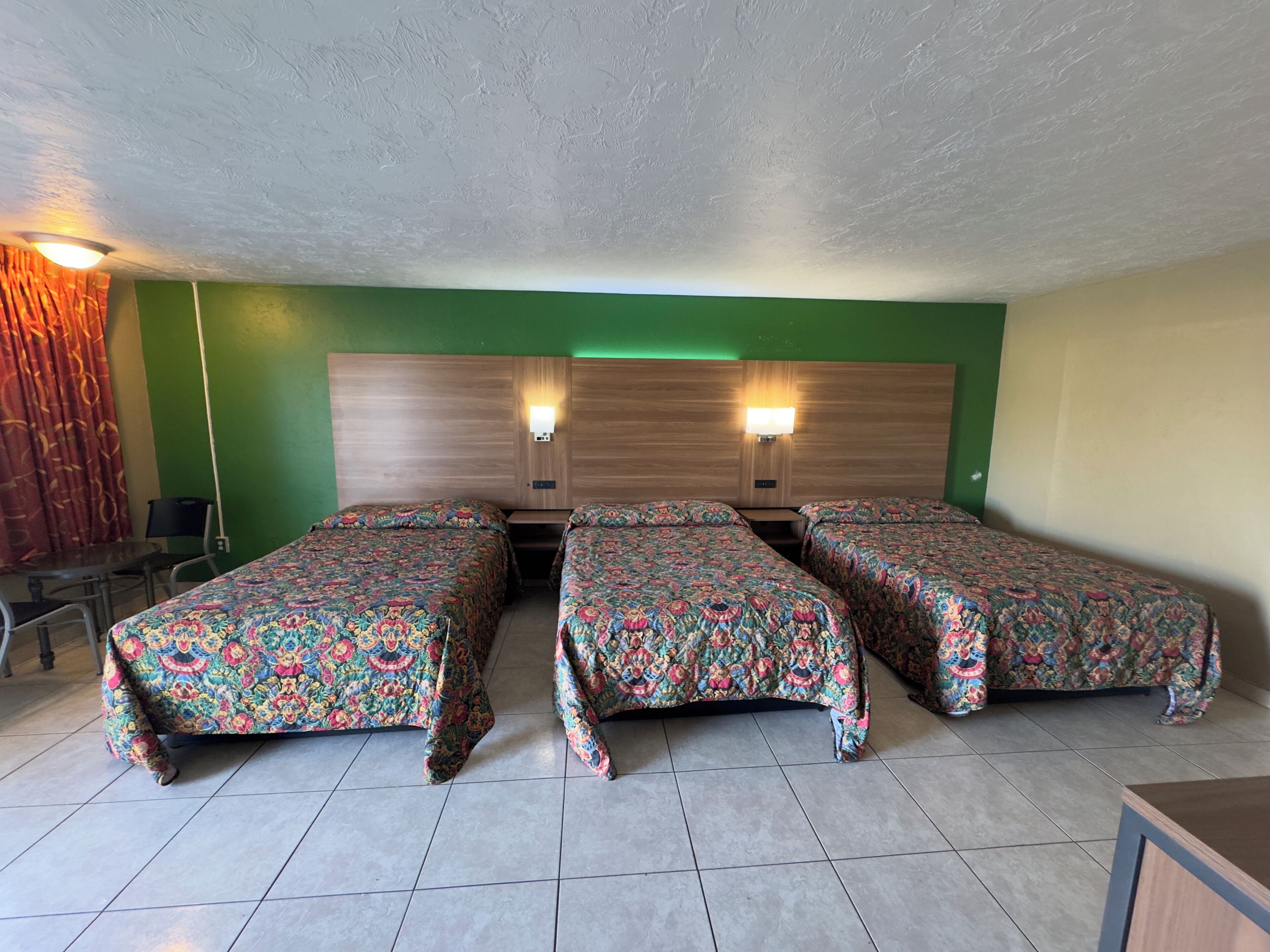 A hotel room with three beds, a wooden headboard, two wall-mounted lamps, a green accent wall, and a tiled floor.