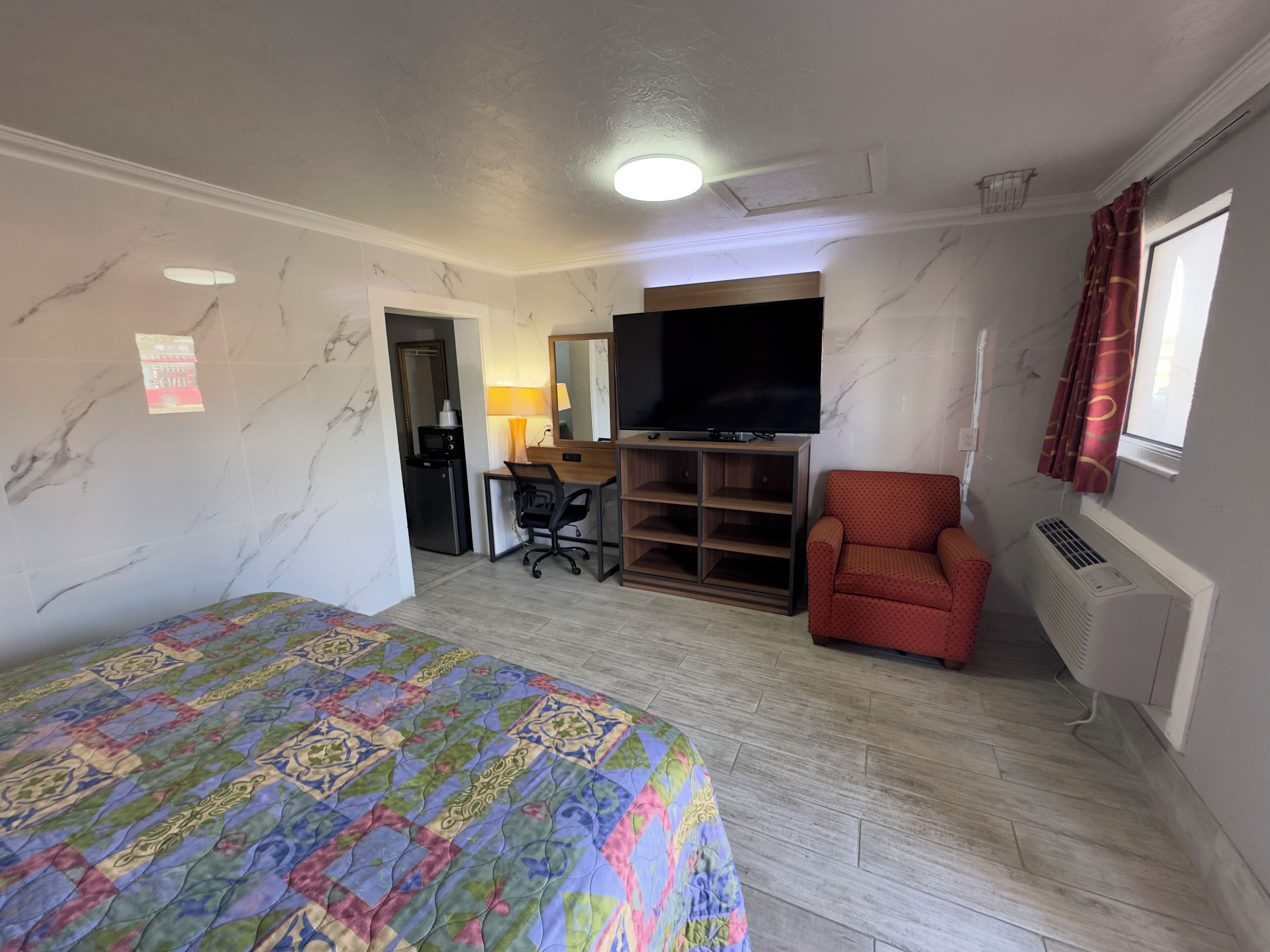 A hotel room with a bed, a desk with a chair, a television, a red armchair, and a window with curtains.