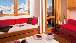 A cozy room with a red sofa, a small table with a plate of food and a mug, and a view of snowy mountains through large windows.