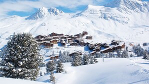 A snow-covered mountain resort with chalet-style buildings, ski slopes, and a clear blue sky.