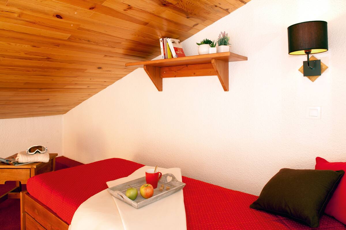 A bedroom with a wooden ceiling, a bed with red bedding, a bedside table with a pair of goggles, a tray with apples and a mug, and a wall-mounted shelf with books and a plant.