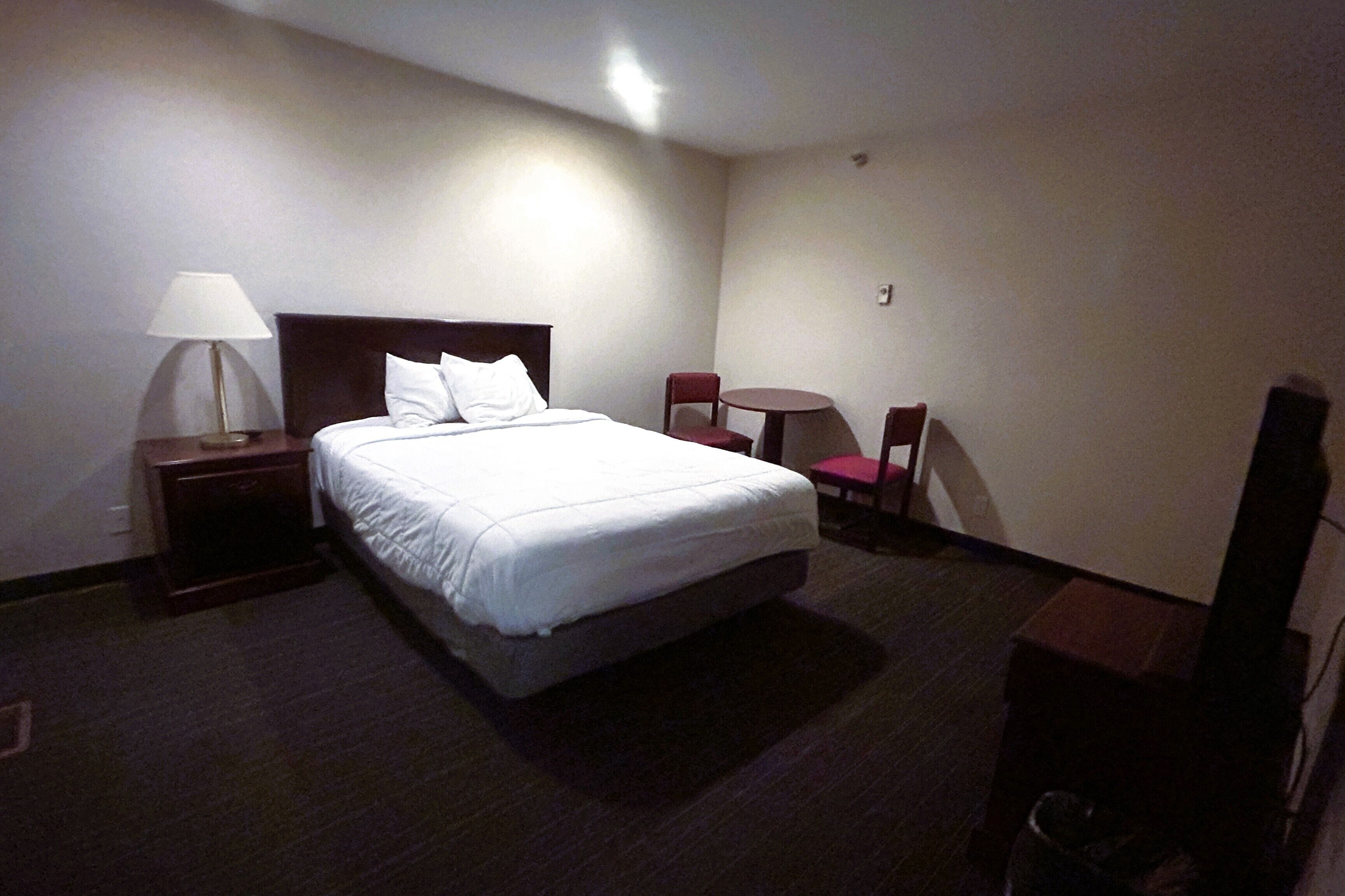 A hotel room with a bed, a nightstand, a lamp, a small table, two chairs, and a television.