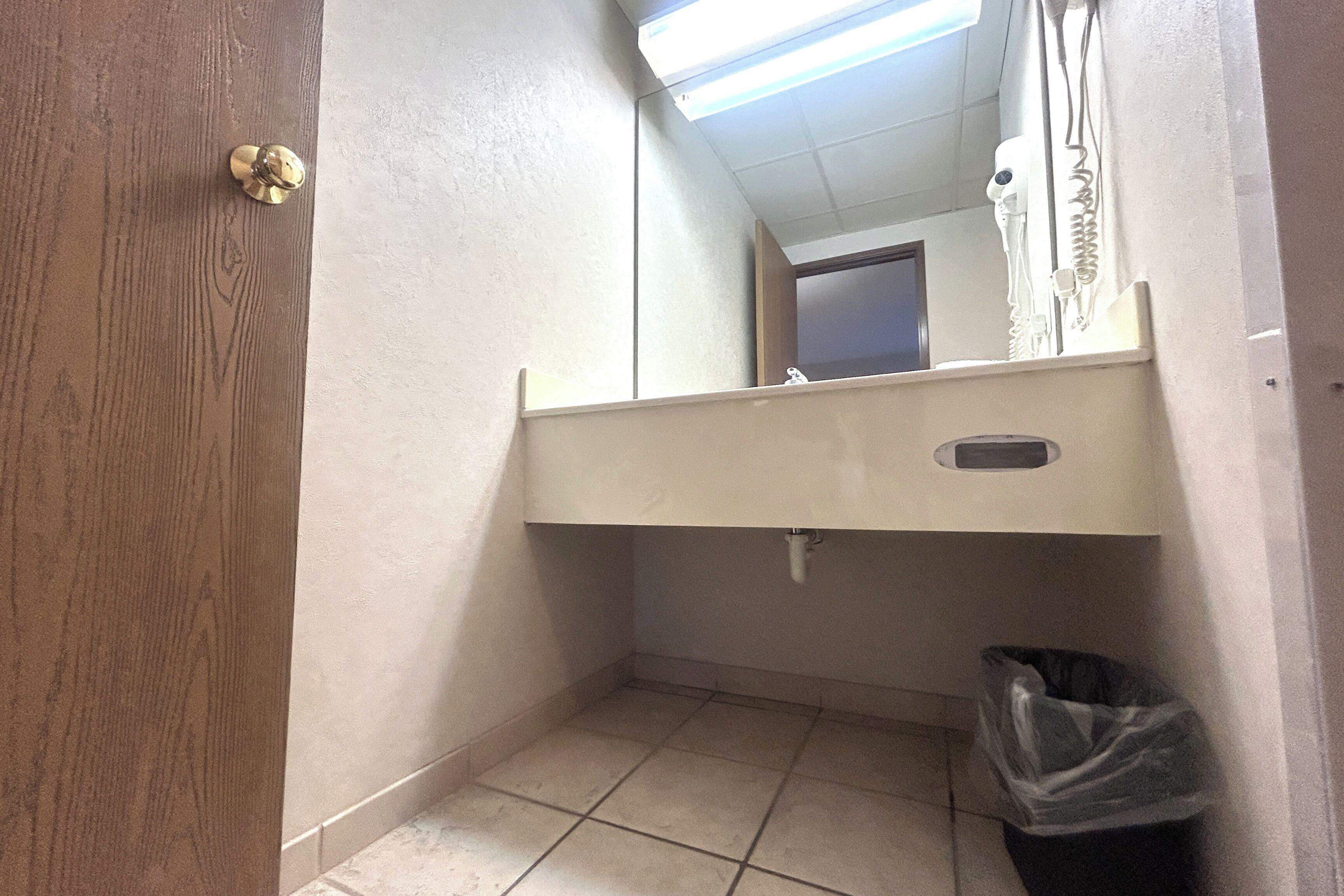A bathroom with a wooden door, a mirror, a sink, and a trash bin.
