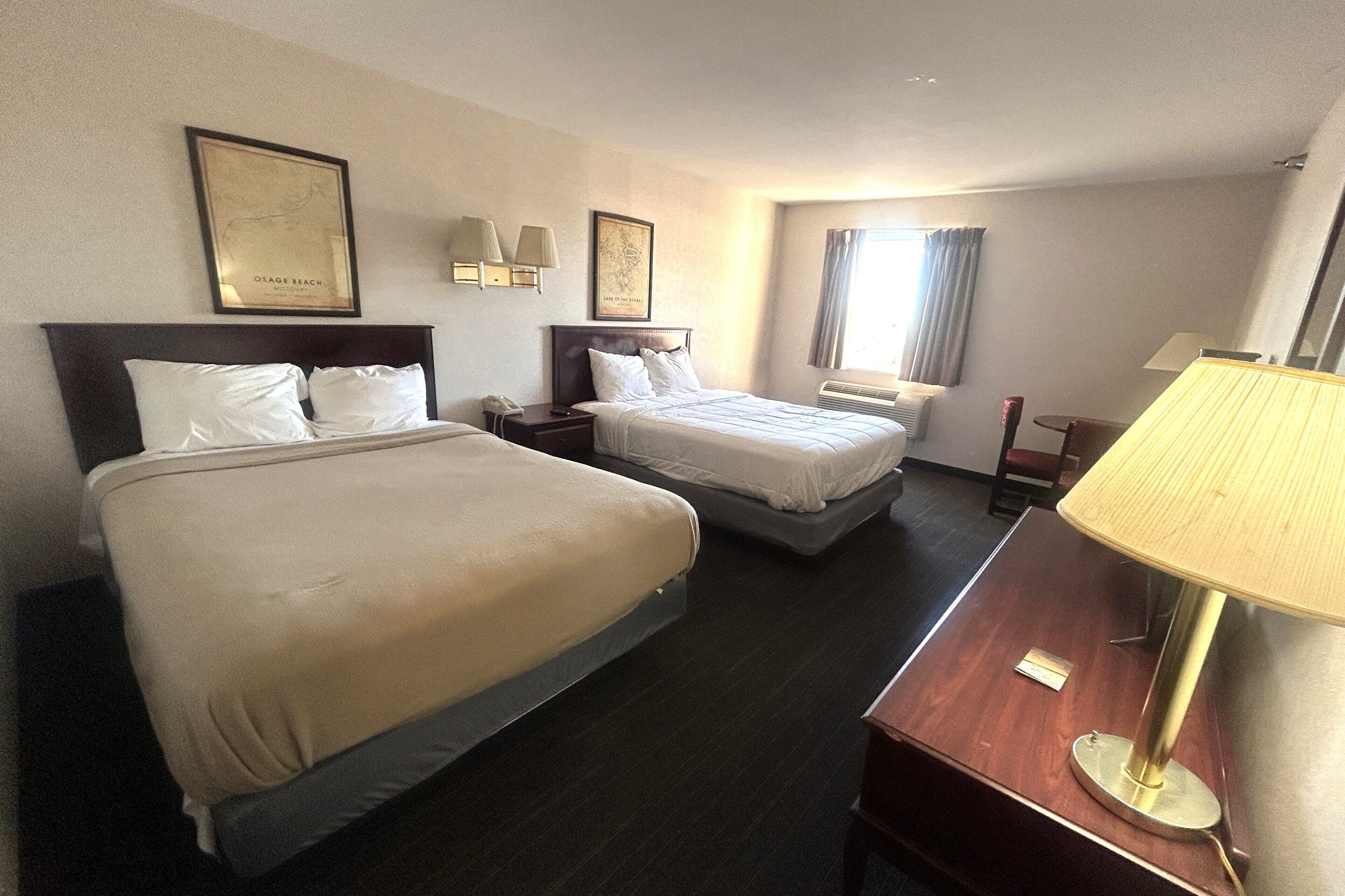A hotel room with two beds, a desk, a lamp, and framed pictures on the walls.