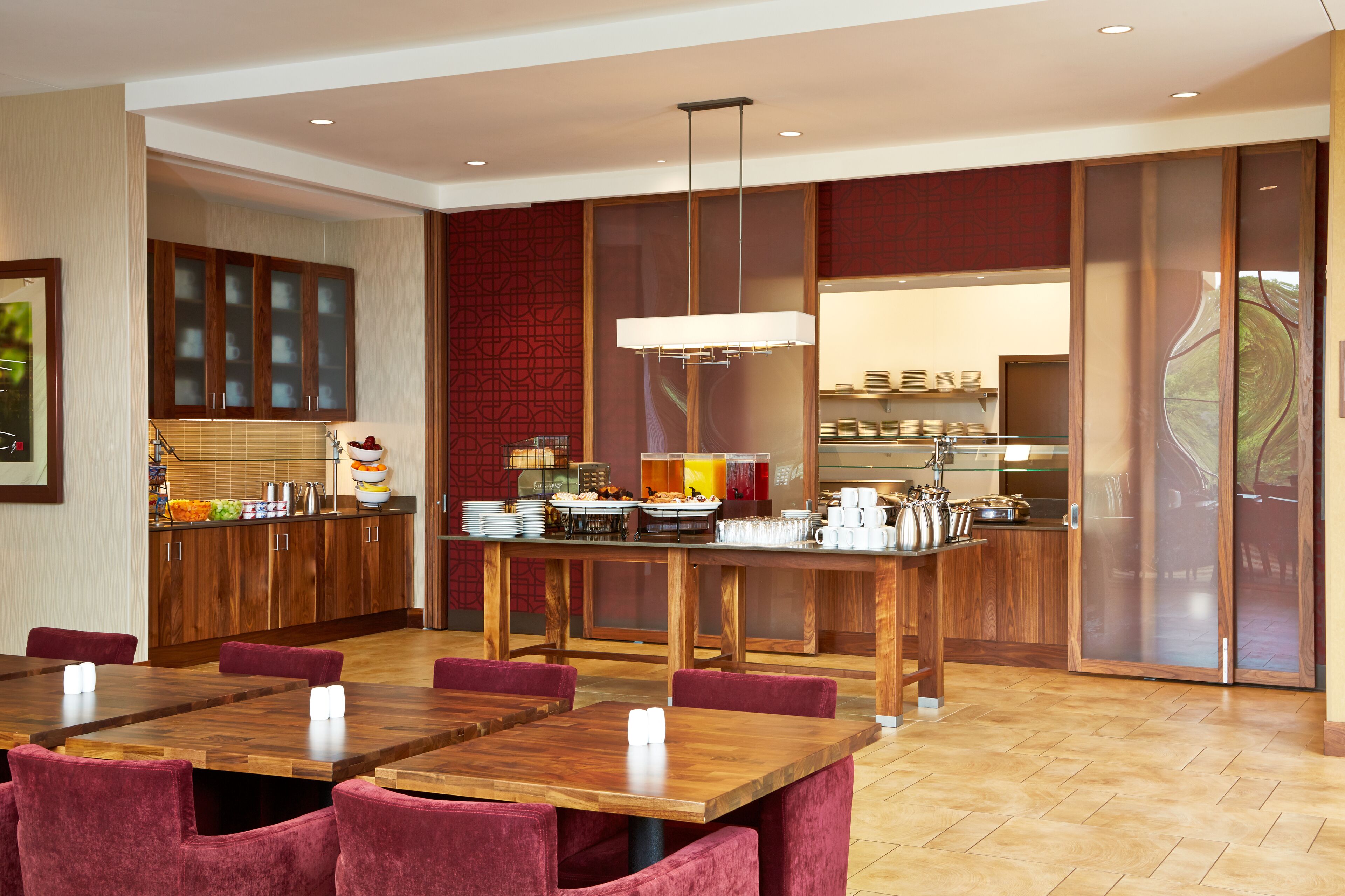 Un comedor moderno con mesas y sillas de madera, una estación de servicio central y una cocina al fondo.