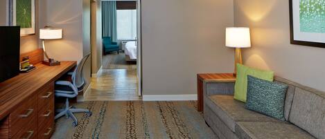 A hotel room with a wooden desk, a sofa with cushions, a patterned carpet, and a hallway leading to another room.