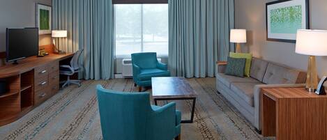 Habitación de hotel con escritorio de madera, televisor, sillón azul, sofá, mesa de centro y ventana grande con cortinas.