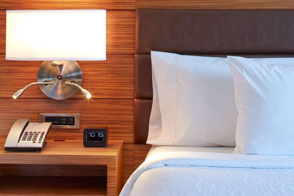 A hotel room with a wooden headboard, a bedside lamp, a clock, a telephone, and a painting on the wall.