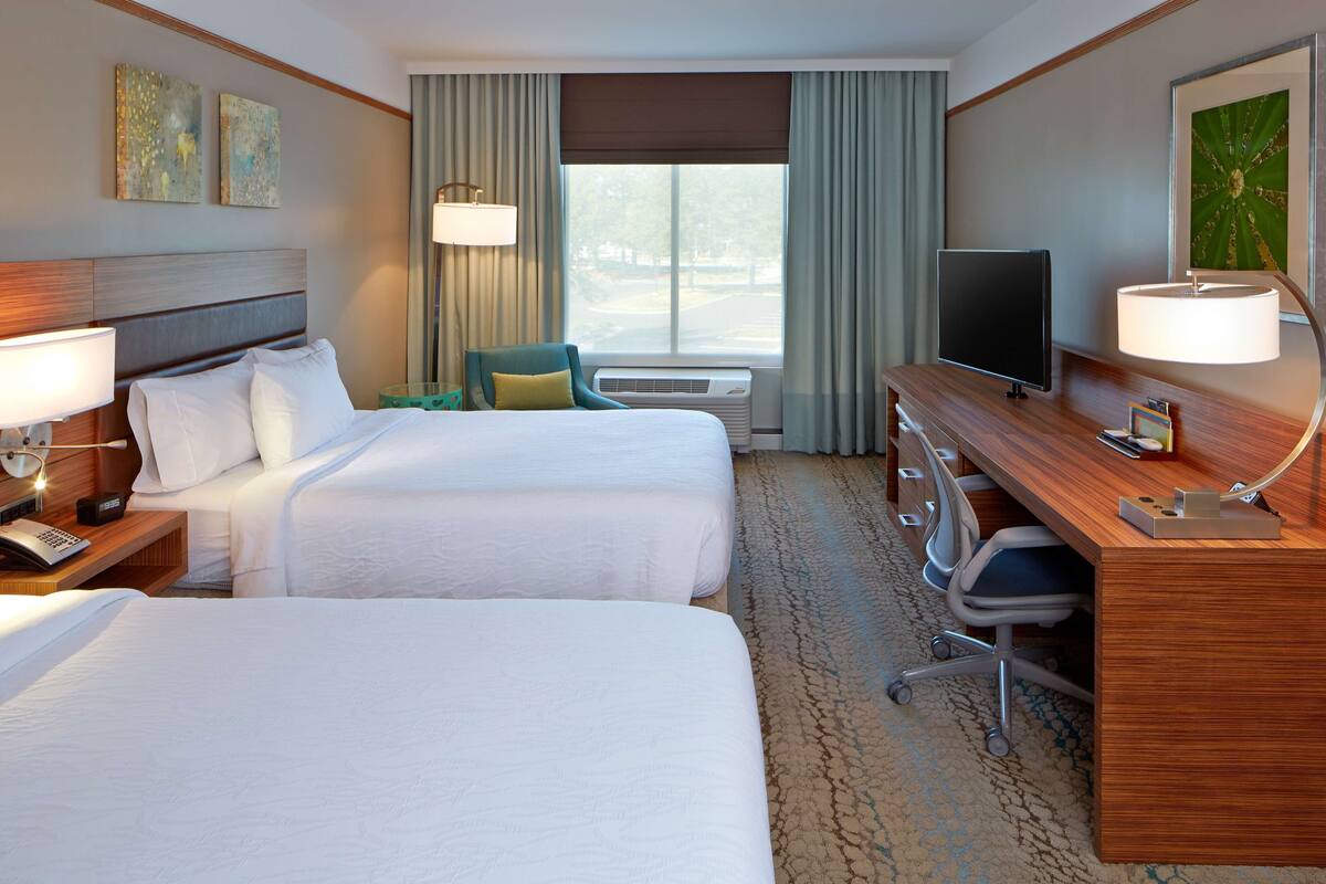 A hotel room with two beds, a desk, a television, and a window with curtains.