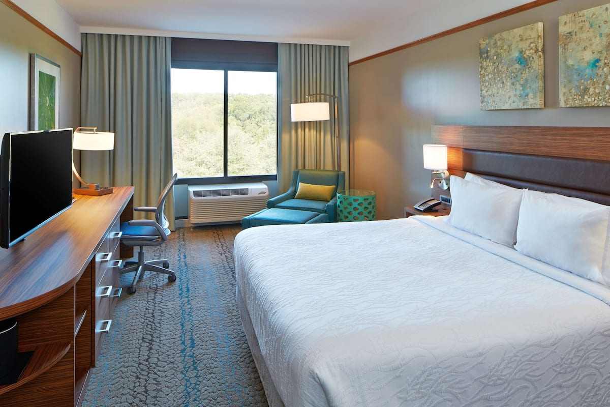 A hotel room with a large bed, a desk with a TV, a chair, a window with curtains, and artwork on the wall.