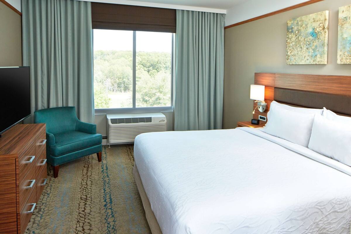 A hotel room with a bed, a television, a chair, a window with curtains, and artwork on the wall.