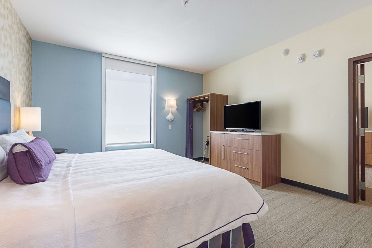 Une chambre d’hôtel avec un lit, un téléviseur posé sur une commode en bois, une fenêtre donnant sur un paysage agréable et un luminaire fixé au mur.