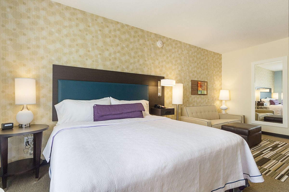 Une chambre d’hôtel avec un grand lit, deux tables de chevet, une petite table, une chaise, un canapé, un miroir et un papier peint à motifs.