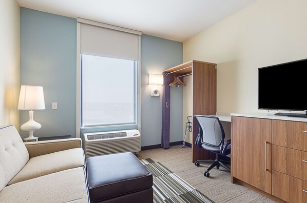 A hotel room with a sofa, a desk with a computer, a television, a window with blinds, a lamp, and a heater.